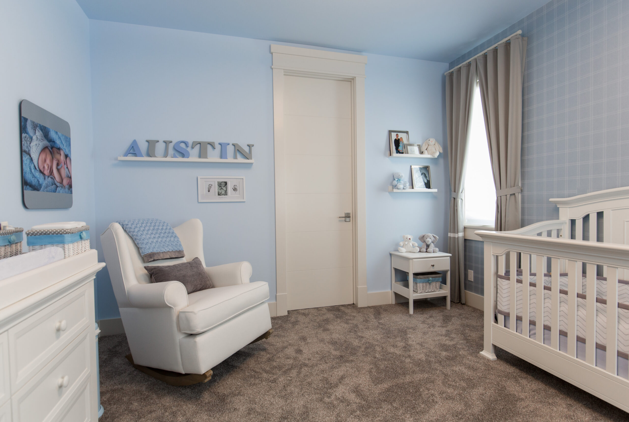 A babies room decorated with light blue walls, striped wallpaper, baby decor, an off-white crib, change table and rocking chair.