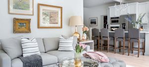 An elegant living room with a grey sofa, coffee table, and a gallery wall of framed artwork. The kitchen is visible in the background with a breakfast bar and chairs.