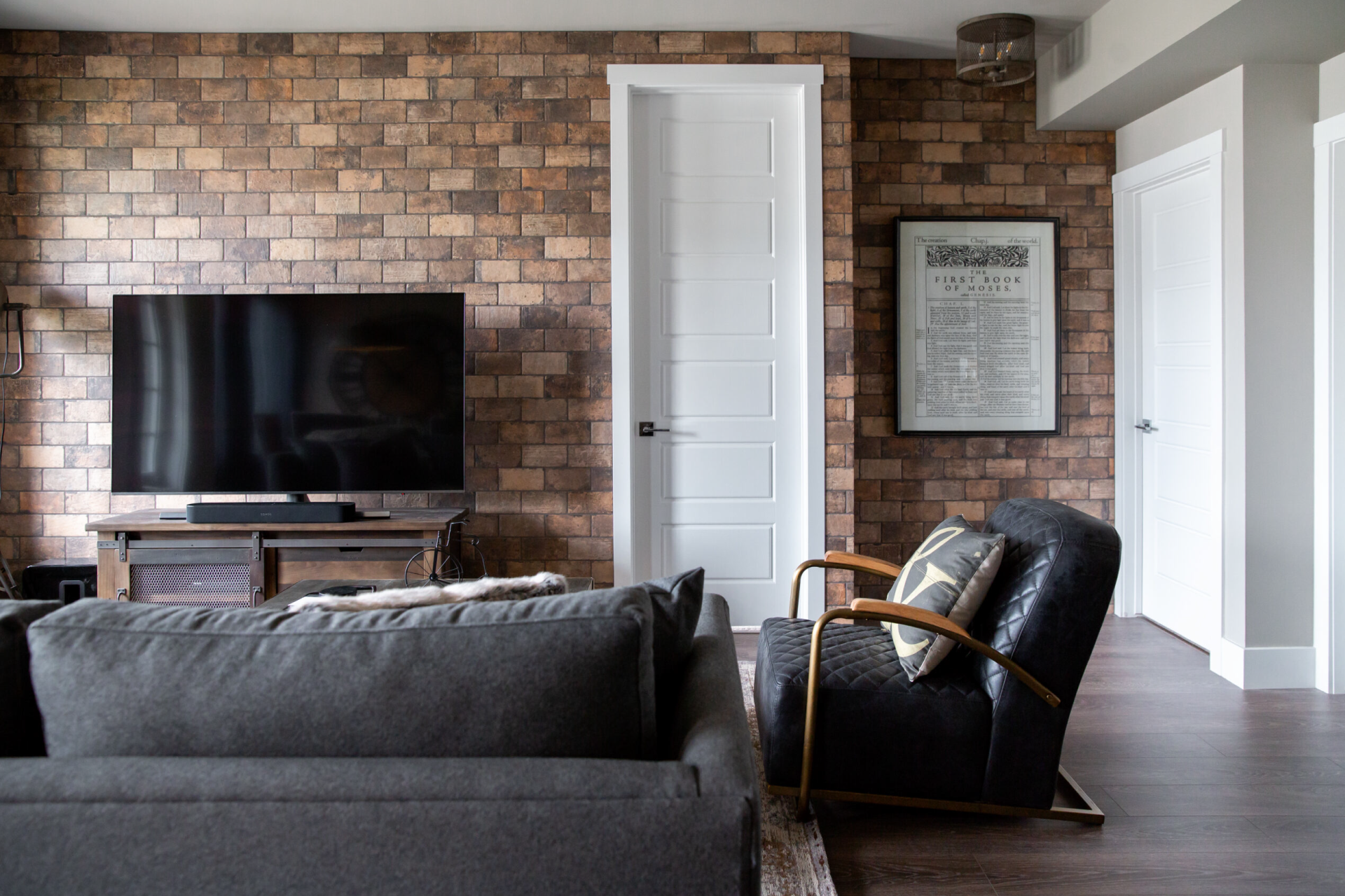 A modern living room with a brick accent wall. A large flat-screen TV is mounted on the wall, and a leather armchair with gold accents sits in front of it.