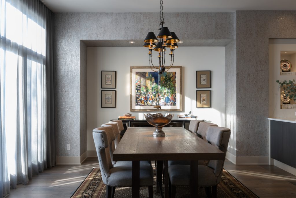 A refined dining room with soft light filtering through grey sheers. A dark wooden table, six upholstered chairs, and a chandelier. It has textured walls with pretty artwork and a modern sideboard located in an alcove at the end of the room.