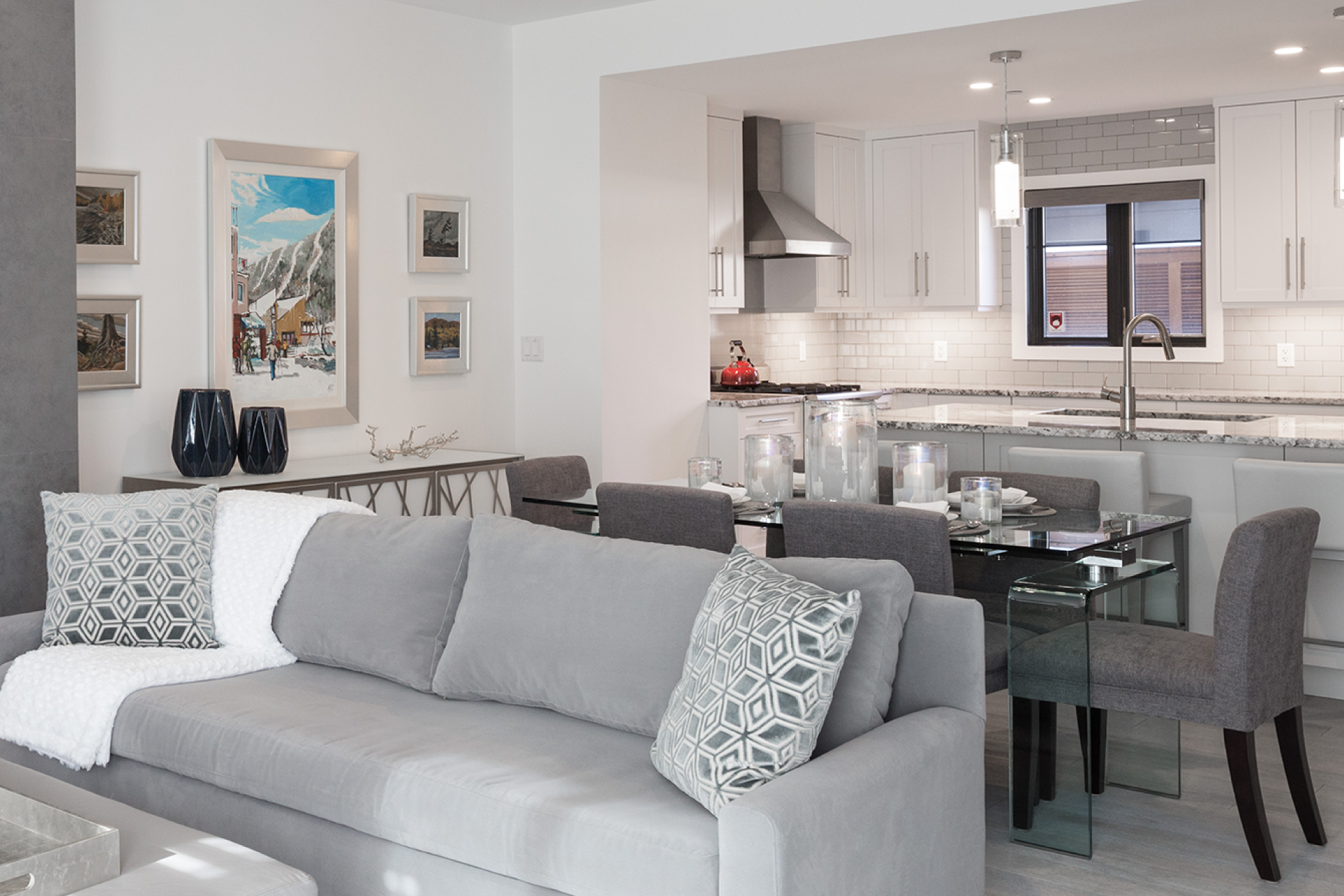 A modern, open-concept living room and kitchen. A gray sofa with pillows sits in the living area, while a dining table with chairs is set up in the kitchen area.