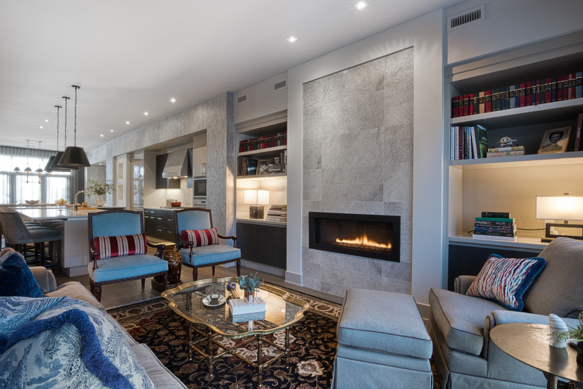 A large modern gas fireplace with grey tile in an open space living room. The kitchen is visible in the background with large island and bar chairs.