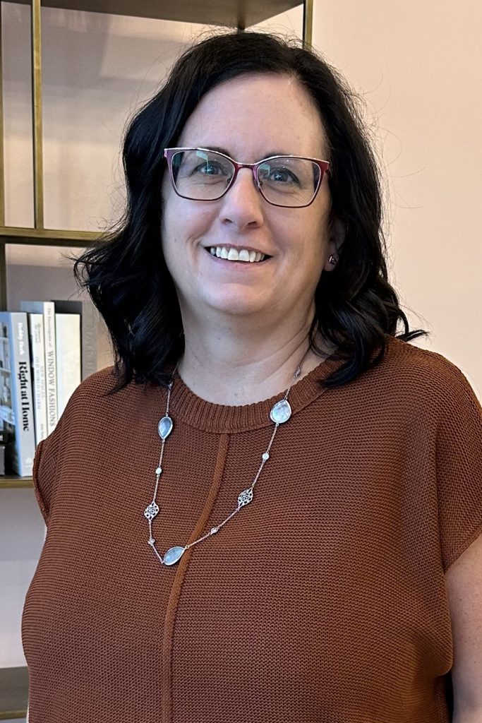 A professional headshot of a woman in business attire smiling and standing in front of a neutral office background.